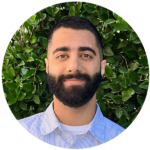 A portrait of a man with a beard smiling, wearing a blue striped shirt, standing in front of a leafy green hedge background in Los Angeles.