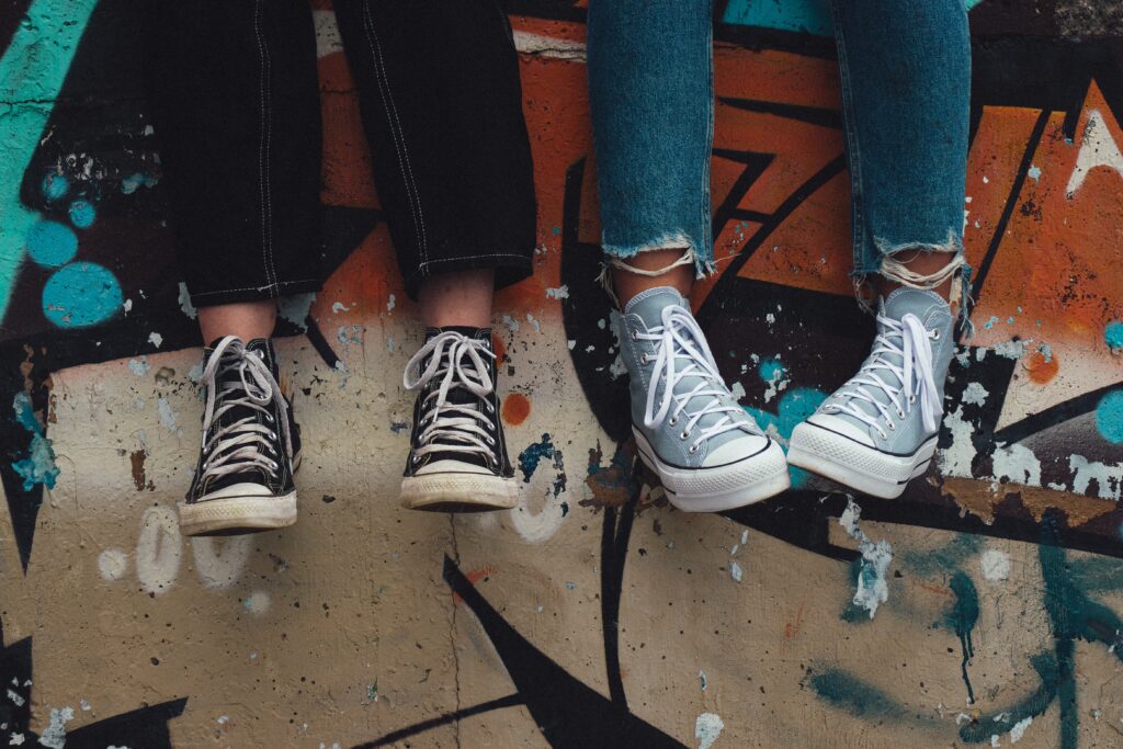 Two people sitting on a graffiti-covered ledge in LA, showing only their legs and sneakers, one wearing black and the other white sneakers.