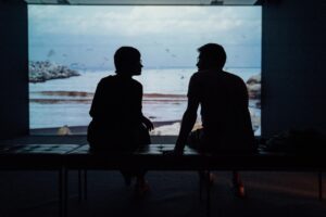 Two people sitting and talking in a dim room, silhouetted against a large screen displaying a serene Los Angeles beach scene.