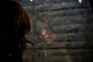 Woman seen in reflection, looking out a dirty window. Her expression is thoughtful, with a blurry wooden background. She is a teen considering therapy.