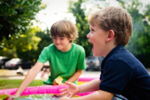 Two teen boys playing with toys on a picnic blanket outdoors, one excitedly shouting while the other reaches towards the toys.