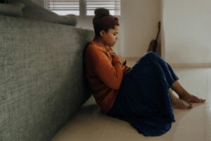 A teen in a brown sweater and blue skirt sits on the floor beside a sofa, hugging her knees and looking thoughtful.