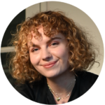 A teen with curly blonde hair and a nose piercing, smiling at the camera in a warmly lit setting.