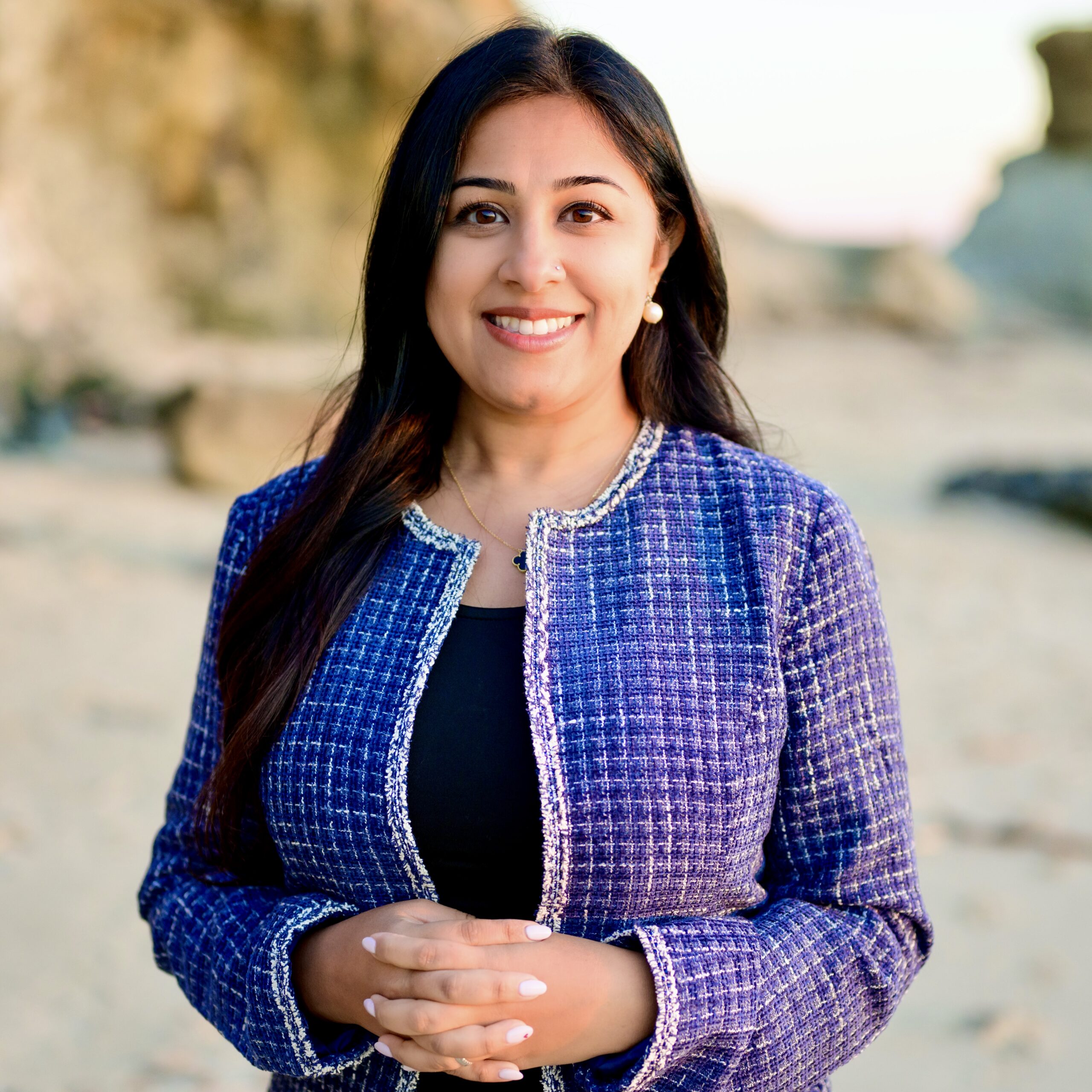 A woman smiling at the beach in LA, wearing a blue tweed jacket and black top, with her hands clasped in front. Rocks and sand are visible in the background.