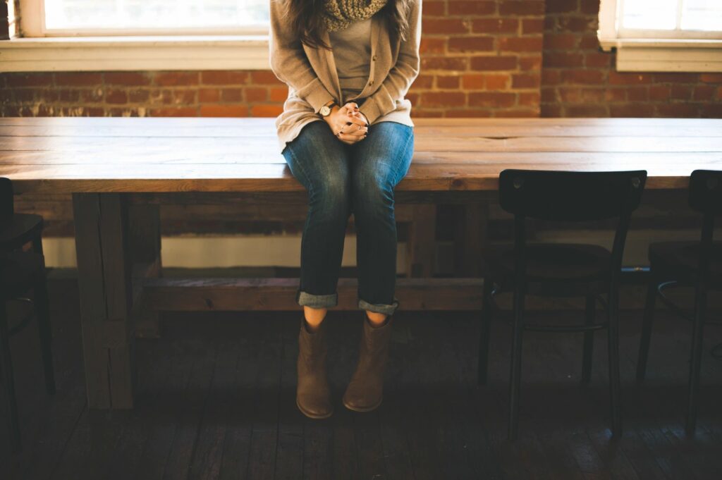 A woman sits on a wooden table in Los Angeles, her hands clasped, wearing jeans and brown boots, with empty chairs and a brick wall in the background.