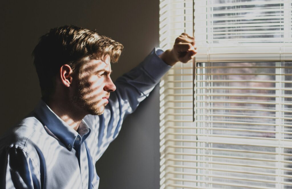 A teen in a blue shirt looks pensively out of a window, his face partially illuminated by sunlight filtering through blinds.
