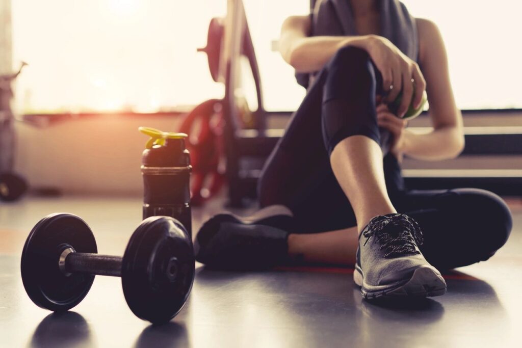 Woman sitting on gym floor next to a dumbbell, holding a water bottle, with focus on her sneaker after family therapy.