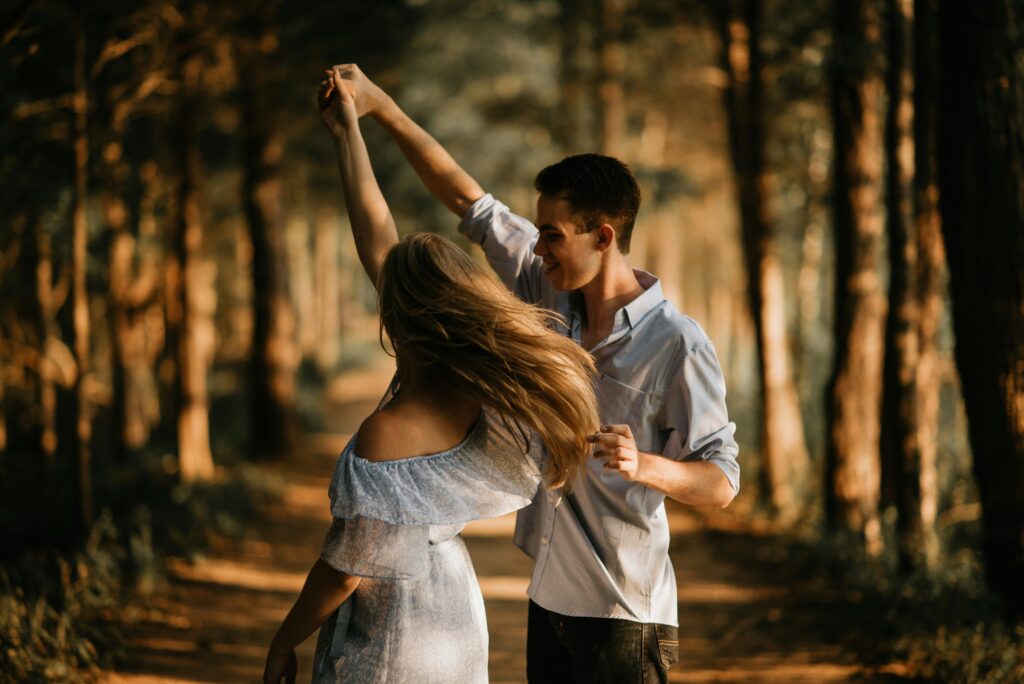 A couple dances on a sunlit forest path in Los Angeles, with the man holding the woman's hand aloft as she twirls in a white dress.