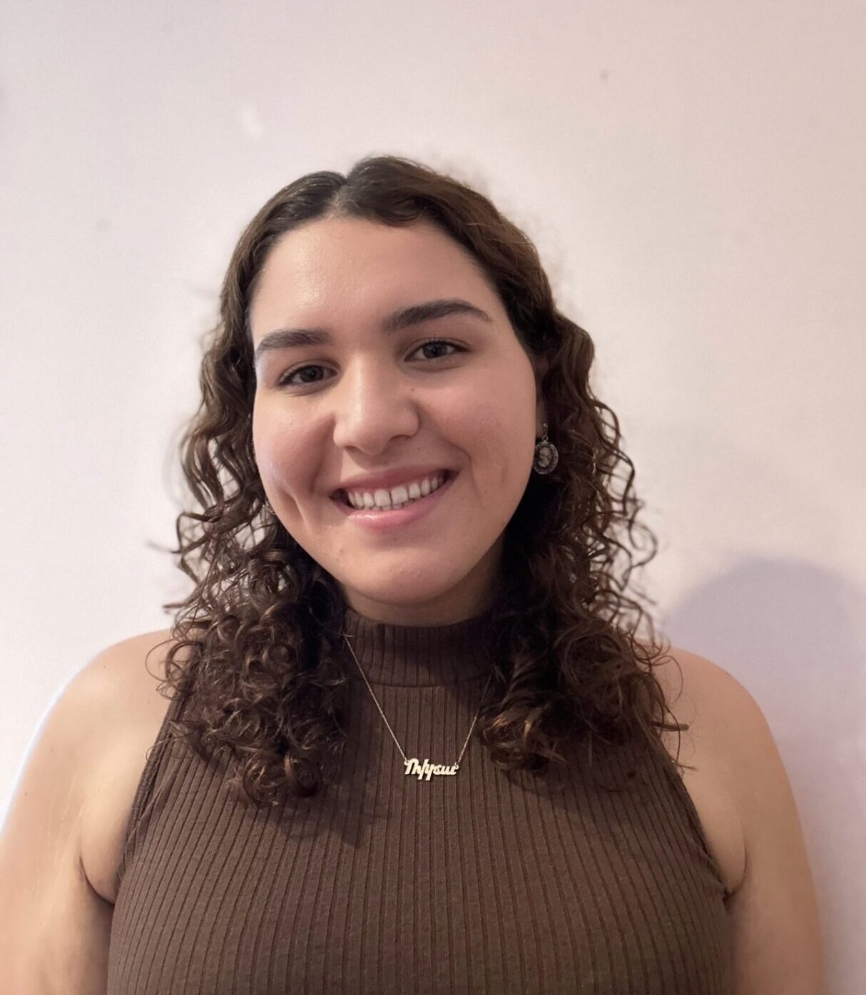 A person with curly hair wearing a brown sleeveless top and a name necklace smiles at the camera against a plain background.