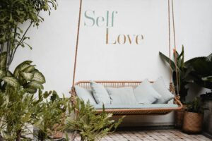 A rattan swing with light blue cushions is surrounded by green plants against a white wall with the words "Self Love" written on it.