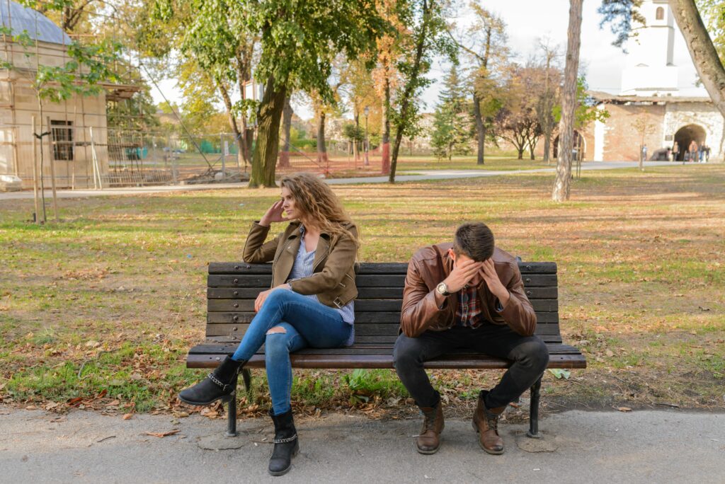 A woman and a man sit on a park bench facing away from each other, both with visible signs of distress.