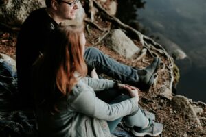 Two people sit on ground outdoors near rocks and roots, looking into the distance. The person in front has long hair and wears a light jacket; the person behind wears glasses and a dark jacket.