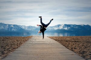 Person performing a one-handed handstand on a wooden path with mountains and a lake in the background.