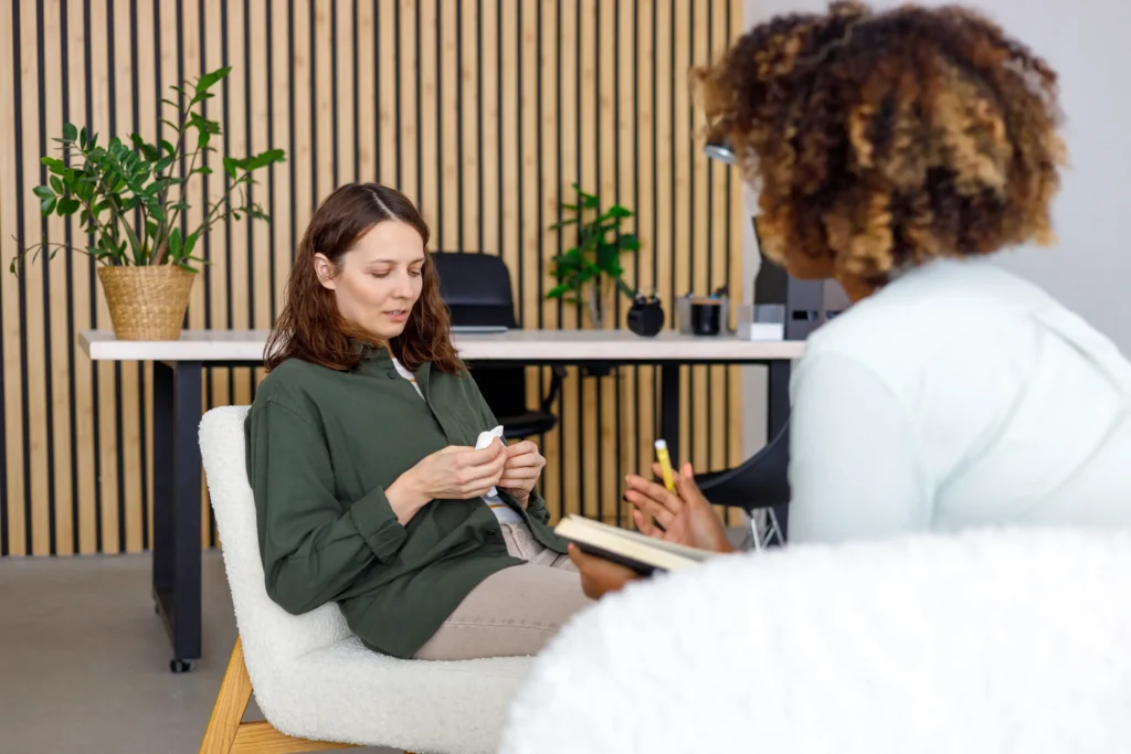 Two people sitting and talking in an office setting. One is holding a notepad, and the other is holding a small object. A desk and plant are in the background.