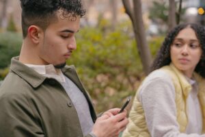 A person with curly hair looks at their phone while another person with curly hair looks at them, sitting in an outdoor setting.