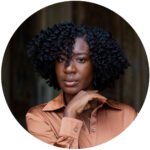 Shamika Anglin A person with curly hair wearing a brown shirt poses with their hand near their chin, looking directly at the camera.