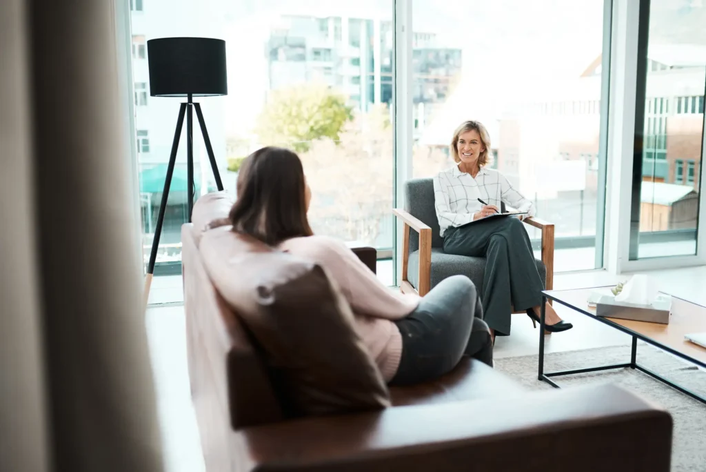 A person sits on a couch talking to another person sitting in a chair, holding a notebook, in a well-lit room with large windows.