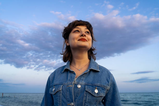 A person in a denim jacket stands on a beach, looking upward, with a cloudy sky and ocean in the background.