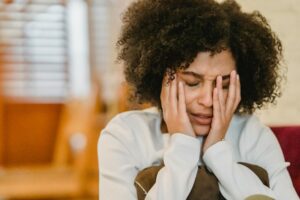 Person with curly hair holding head in hands, appearing upset or stressed, sitting indoors.