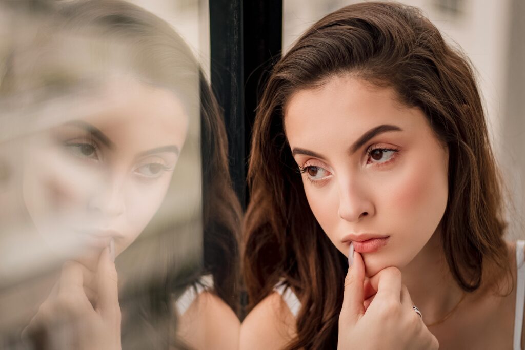 Woman with long hair looks thoughtfully out a window, reflecting softly on the glass.