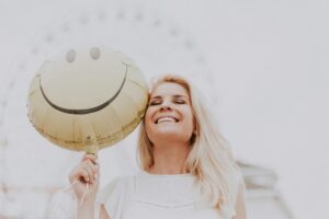 A smiling woman holds a yellow smiley face balloon outdoors. Ferris wheel blurred in the background.