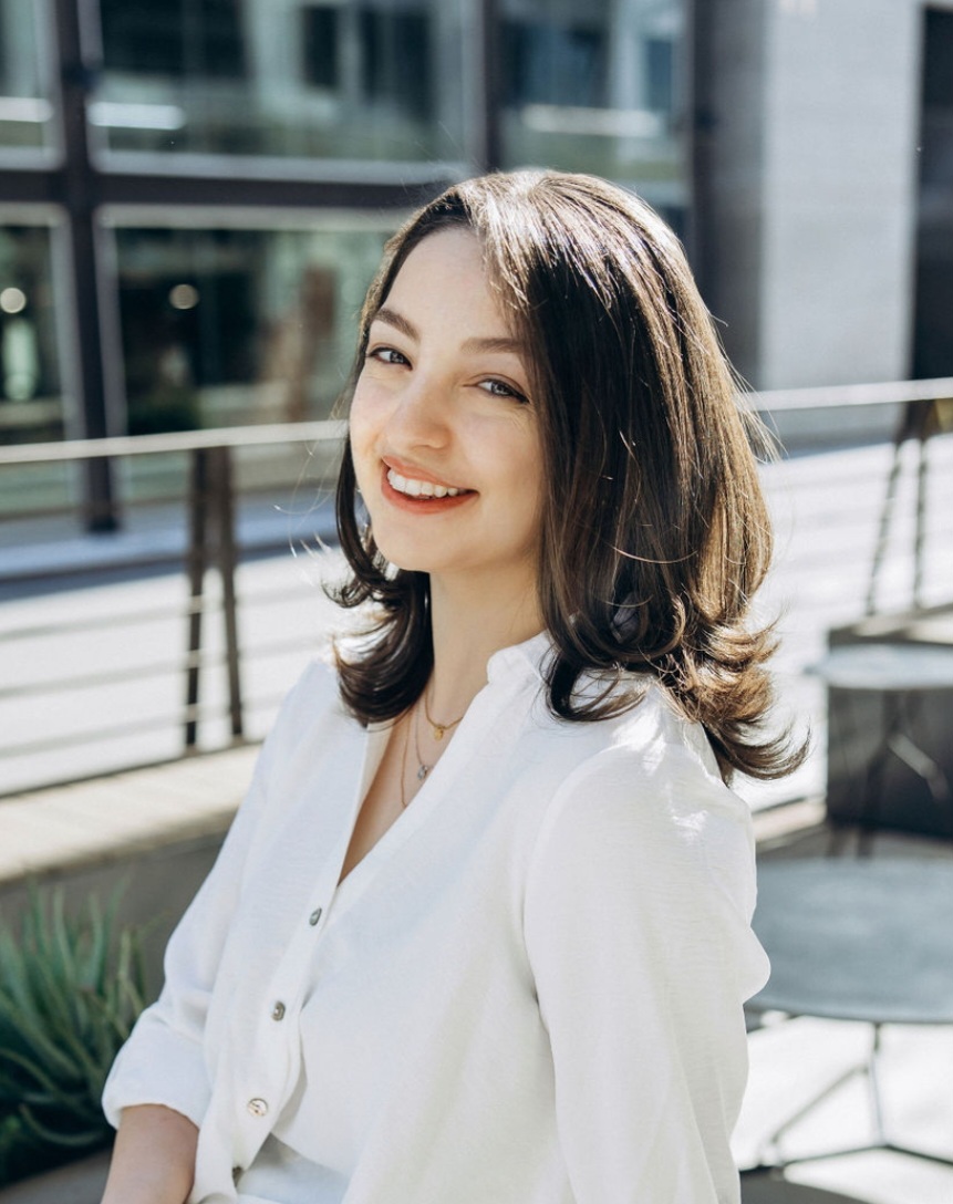 A person with shoulder-length hair smiles while sitting outdoors, wearing a white shirt. Urban background with railing and windows.