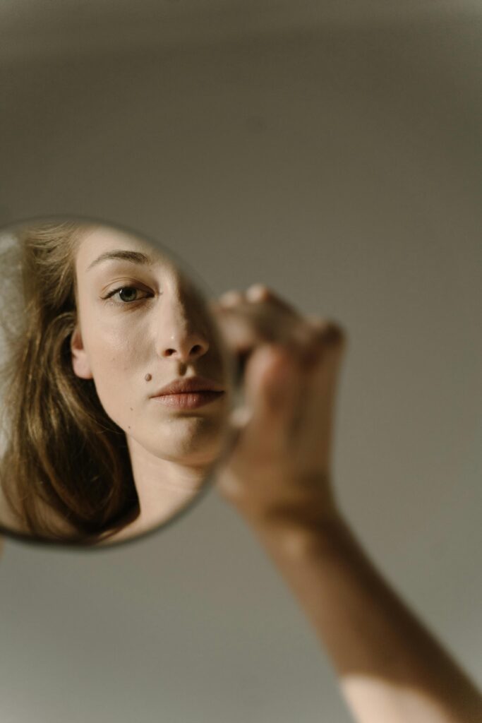 A woman holds a small round mirror reflecting half of her face against a neutral background.