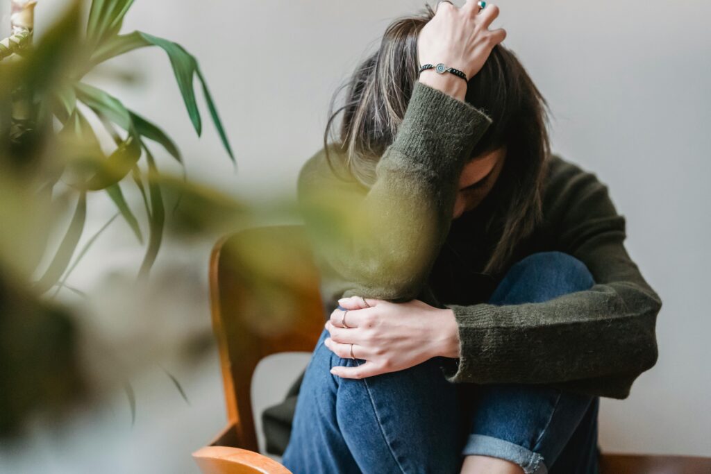 A person sits on a chair, wearing a green sweater and jeans, with their head resting on one arm and holding their knee with the other hand. A plant is partially visible in the foreground.