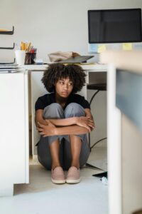 A person sits on the floor under a desk, hugging their knees, with a distant expression. Nearby are office supplies, a cup, and a computer monitor.