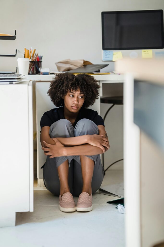 A person sits on the floor under a desk, hugging their knees, with a distant expression. Nearby are office supplies, a cup, and a computer monitor.