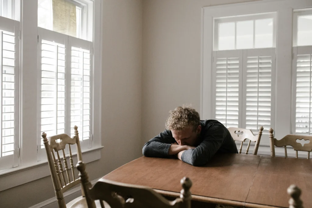 Person sitting at a wooden table, their head resting on folded arms in quiet reflection. The room, ideal for anger management practice, has white walls and shuttered windows, with wooden chairs neatly arranged around the table.