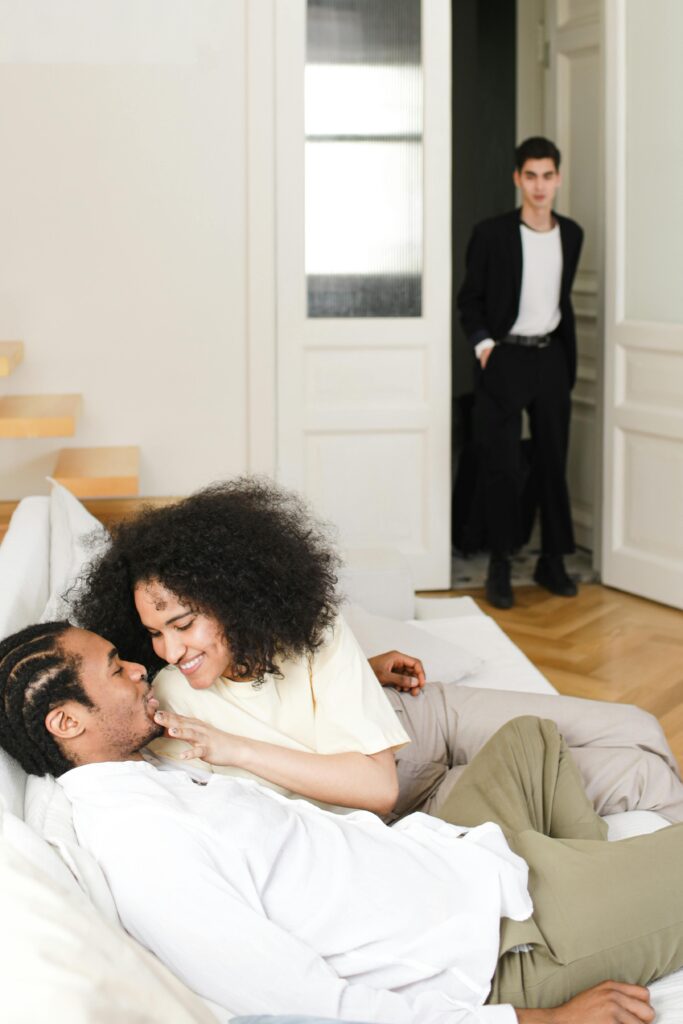 A couple is seated closely on a sofa, smiling at each other. A person in a suit stands in the background, looking towards them from a doorway.