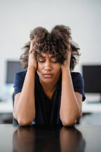 A person sitting at a desk with eyes closed, holding their head in both hands, appearing stressed or overwhelmed.