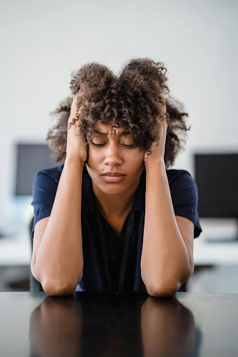A person sitting at a desk with eyes closed, holding their head in both hands, appearing stressed or overwhelmed.