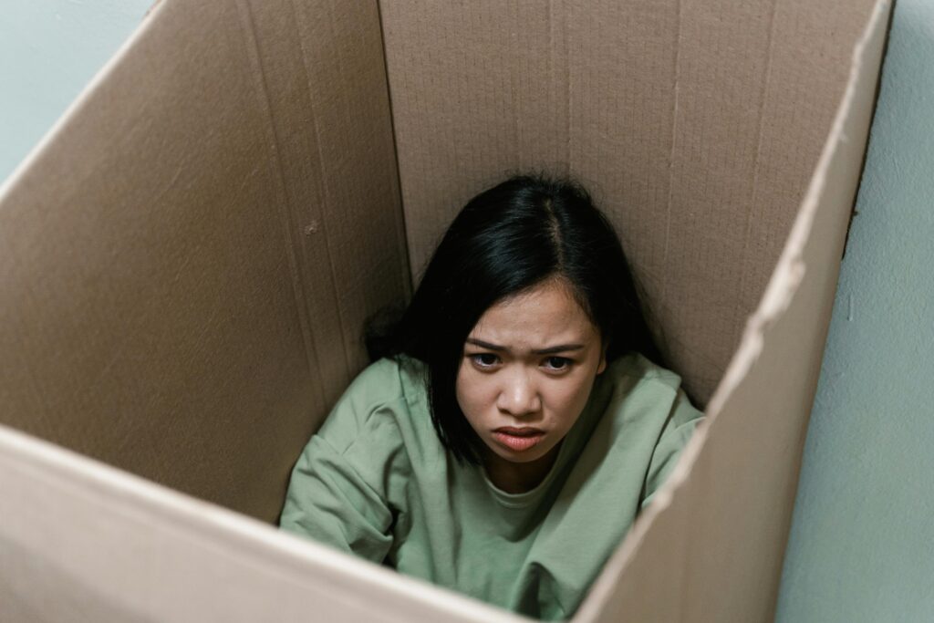 A woman sits inside a large cardboard box, looking up with a concerned expression.