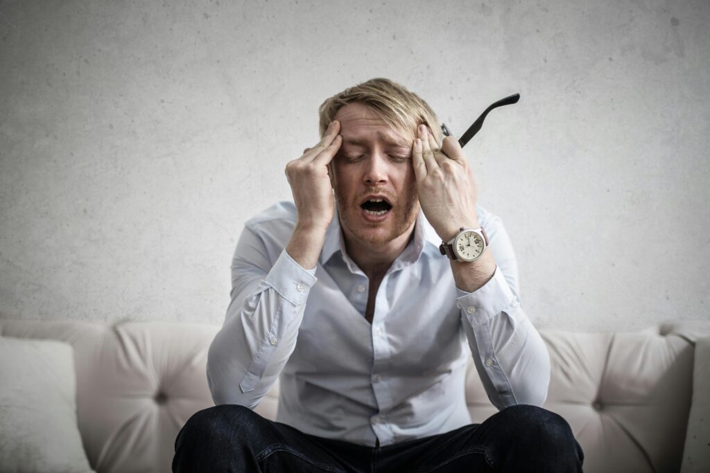 A man sitting on a couch with his eyes closed, touching his temples with both hands, appearing stressed or frustrated.