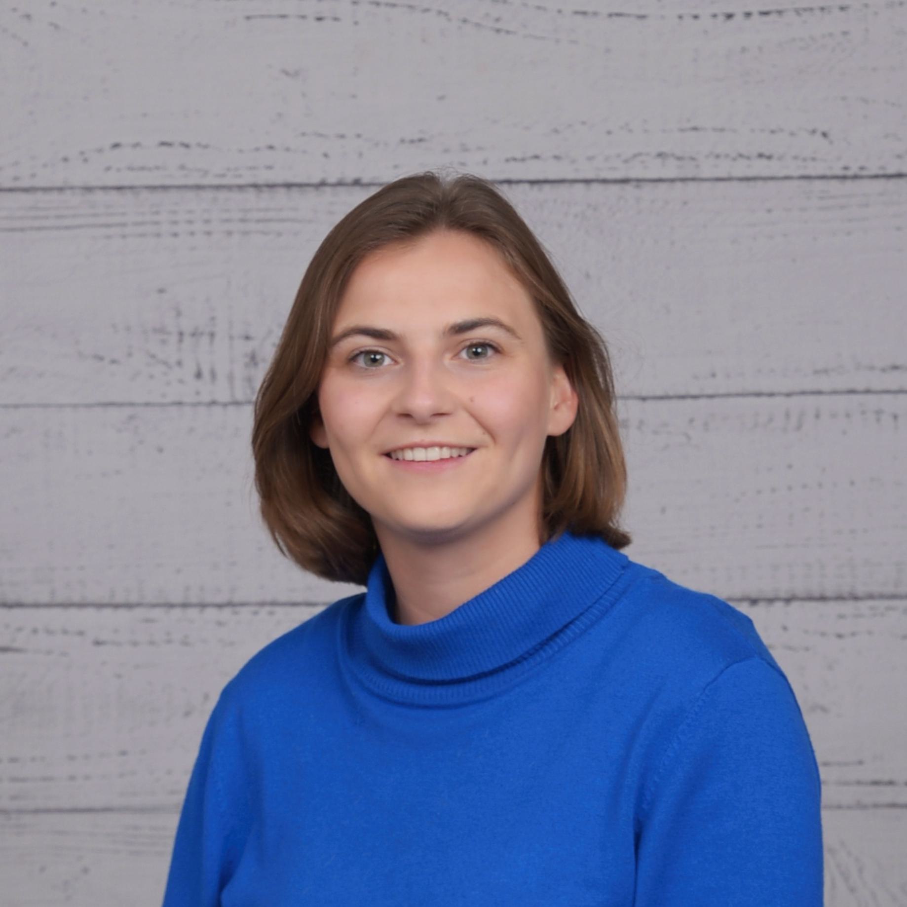 A woman with short brown hair wearing a blue sweater is smiling in front of a light gray wooden panel background.