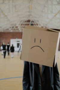 Person wearing a cardboard box with a sad face drawn on it stands in a gymnasium, while other people gather in the background.