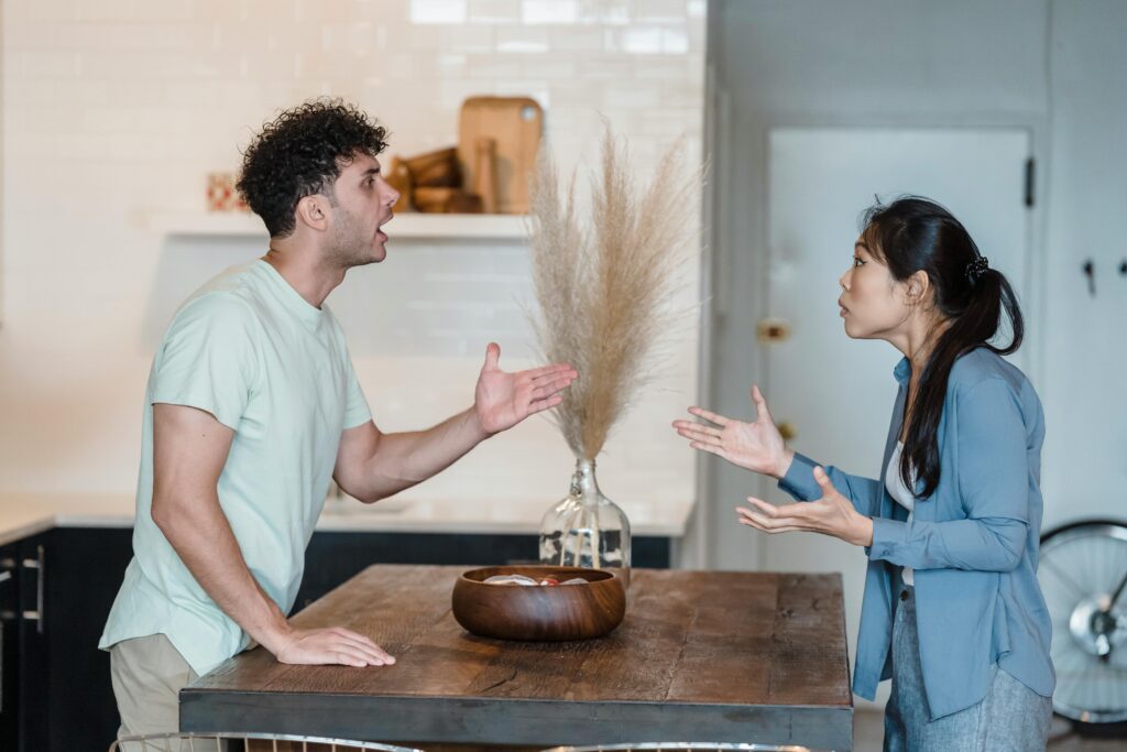 A man and a woman stand on opposite sides of a kitchen table, engaged in a heated argument with expressive gestures.