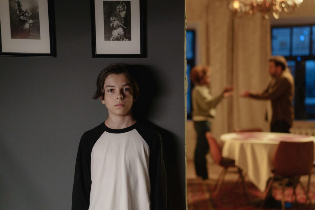 A boy stands alone against a wall, looking at the camera, while two adults argue in the blurred background of a dimly lit room.