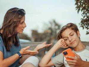 A woman gestures while speaking to a teenager who is looking at their phone and smiling, sitting together outdoors.
