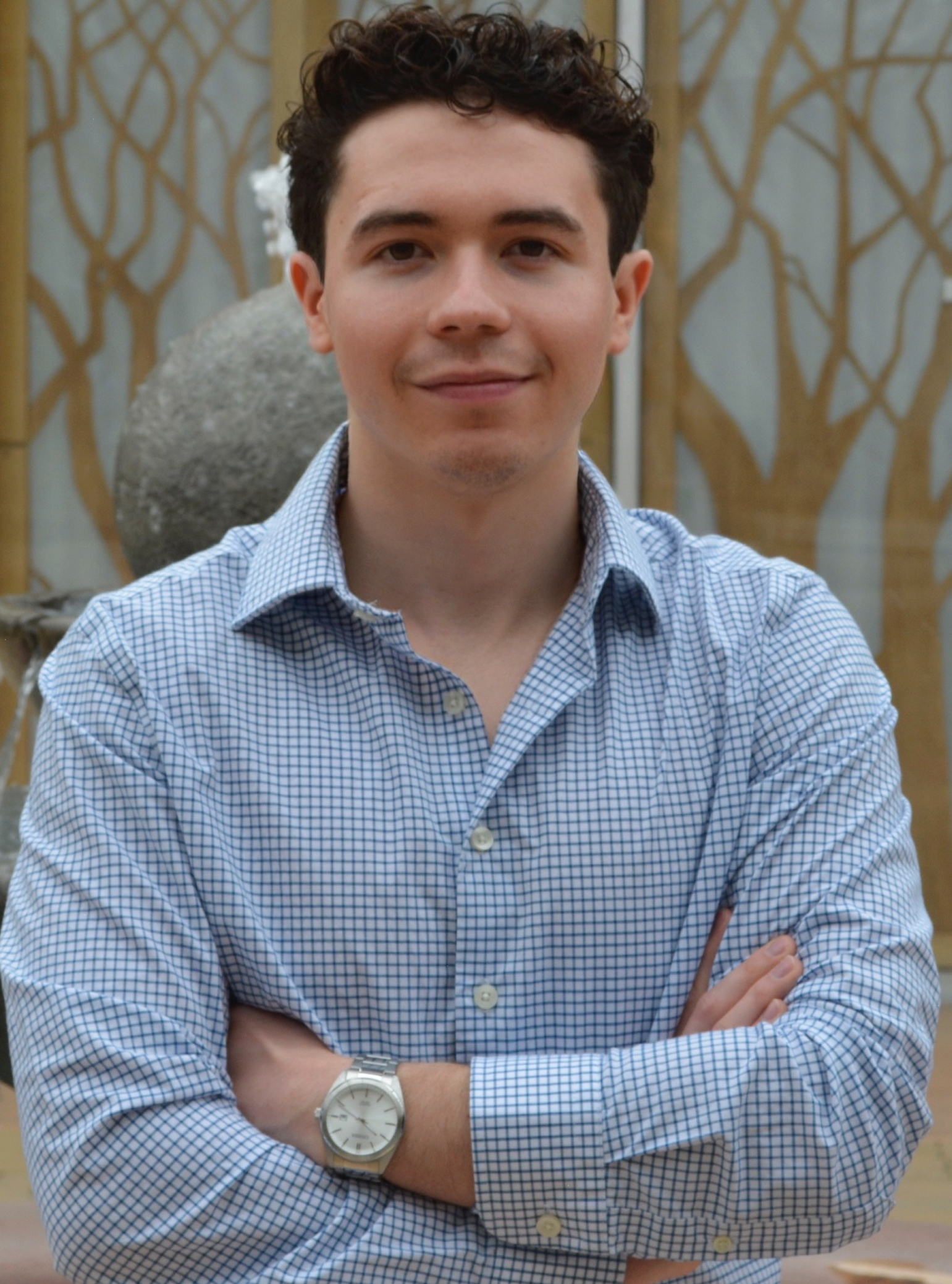 A person with short curly hair, wearing a light blue checkered shirt and a wristwatch, stands with arms crossed in front of a patterned background.