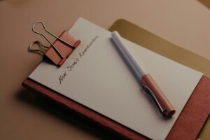 A white pen rests on a clipboard holding a sheet of paper titled "New Year's Resolutions" secured by a pink binder clip.
