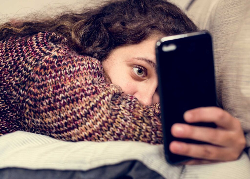 A young woman lying down and scrolling on her phone, illustrating the mental health impact of late-night phone addiction.