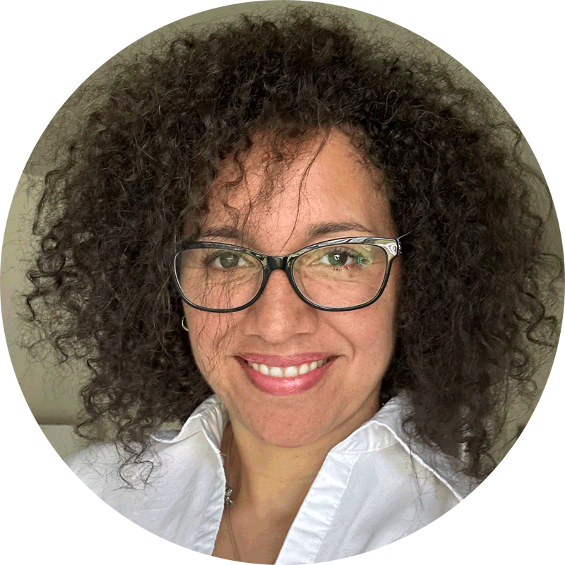 Woman with curly dark hair and glasses, wearing a white collared shirt, smiling at the camera against a neutral background.