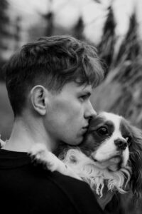 A young man in profile holds and rests his face against a small dog, both looking in the same direction; black and white outdoor setting.