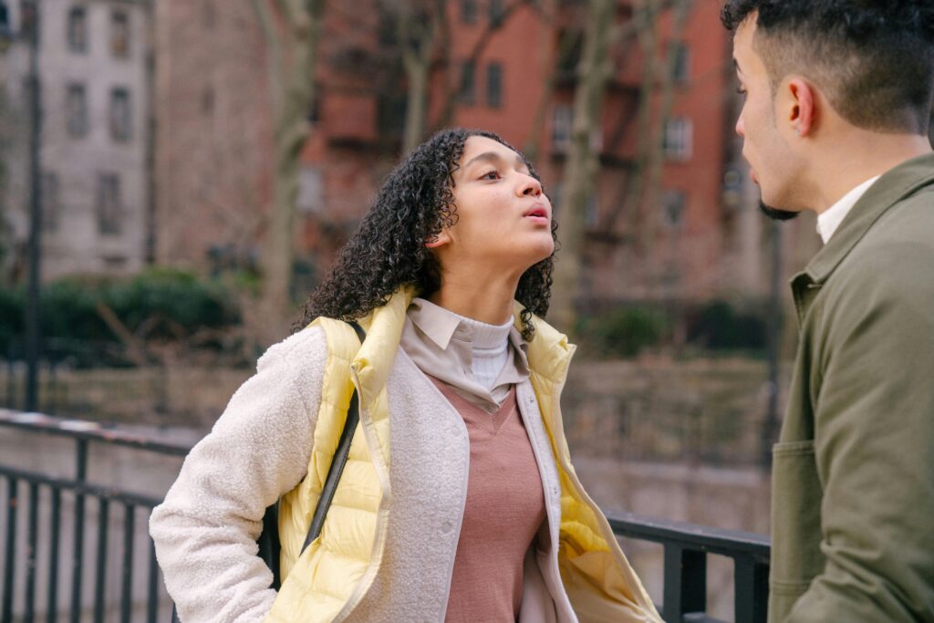 A woman with curly hair wearing a yellow vest speaks emphatically to a man outdoors near a railing with blurred trees and buildings in the background.