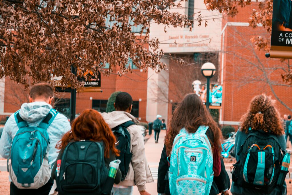 Five students wearing backpacks walk outdoors on a college campus, with buildings and trees visible in the background.