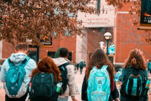 Five students wearing backpacks walk outdoors on a college campus, with buildings and trees visible in the background.