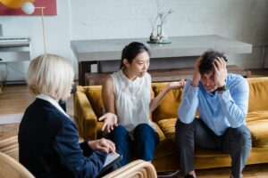 A woman gestures while talking to a therapist, as a man sits beside her with his head in his hands on a yellow couch.